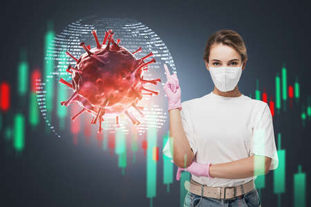 Beautiful young businesswoman in protective mask and rubber gloves standing over blue background with planet hologram, graphs and double exposure of red coronavirus. Toned imageの写真素材