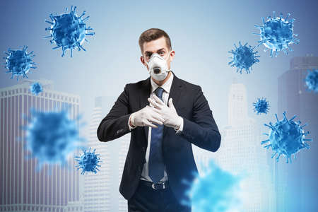 Young European businessman standing in protective mask and rubber gloves in blurry city with double exposure of coronavirus. Toned imageの写真素材