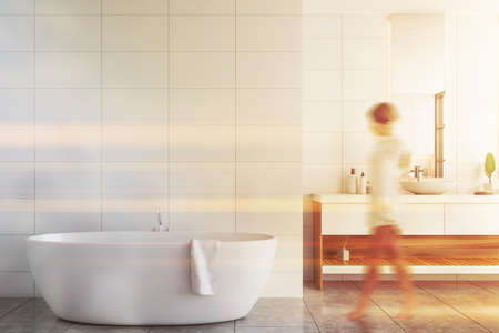 Blurry young woman walking in modern bathroom with white walls, tiled floor, comfortable bathtub and double sink. Toned imageの写真素材