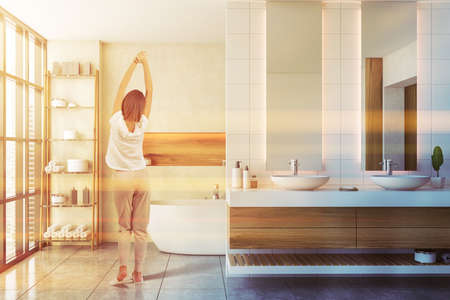 Rear view of young woman standing in stylish bathroom with white walls, tiled floor, bathtub and double sink. Toned imageの写真素材