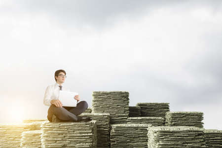 Young European businessman with laptop sitting on stacks of dollars over blue sky background. Concept of investment and wealth.の写真素材