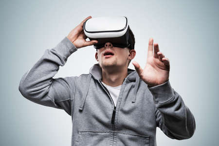 Portrait of young man in casual clothes using VR glasses over gray background. Concept of hi tech...
