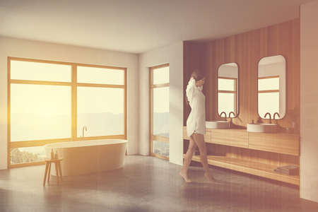 Beautiful young European woman walking in modern bathroom corner with white and wooden walls, concrete floor, double sink and comfortable bathtub. Toned image double exposureの写真素材