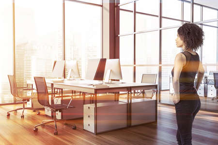 Beautiful young African American businesswoman standing in modern open space office with gray walls, wooden floor and white computer tables. Toned imageの写真素材