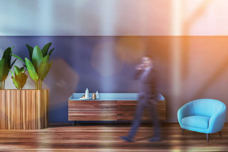 Blurry young man walking in modern living room with white walls, wooden floor and blue armchair....