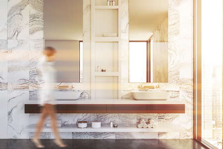 Blurry young woman walking in luxury bathroom with white marble walls, gray floor and double sink with mirrors. Toned imageの写真素材