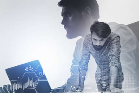 Two serious young businessmen working together with laptop and double exposure of financial chart. Toned imageの写真素材