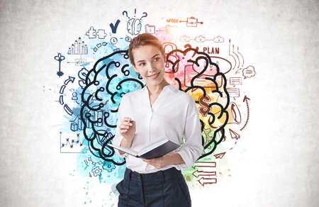 Beautiful young businesswoman with notebook standing near concrete wall with brain drawn on it. Concept of brainstormingの写真素材
