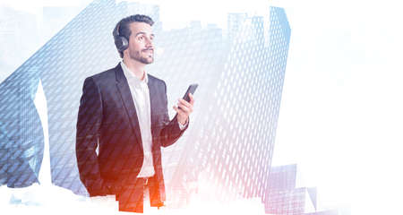 Portrait of young businessman with smartphone and headphones sntanding in blurry city. Concept of...