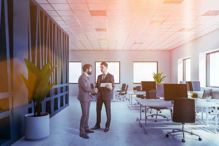 Portrait of two young businessmen standing in modern open space office with white and gray walls and...