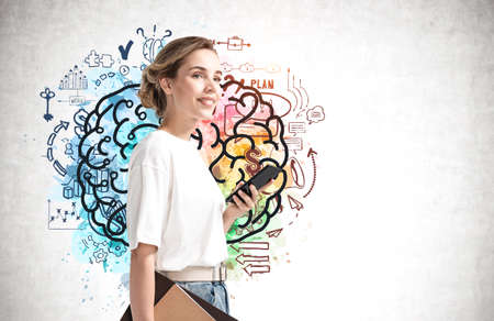 Portrait of smiling young businesswoman with folder and smartphone standing near concrete wall with colorful brain sketch darwin on it.の写真素材