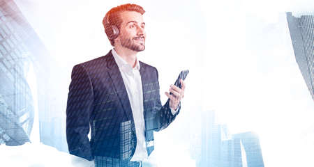 Portrait of young bearded businessman with smartphone and headphones sntanding in blurry city....