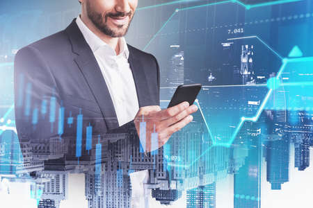 Smiling young businessman using smartphone in blurry night city with double exposure of financial...