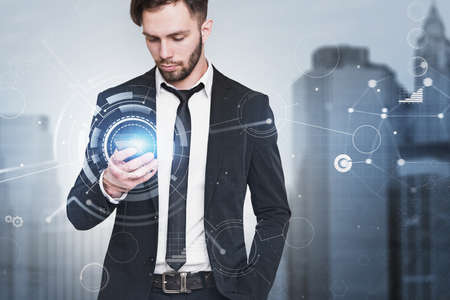 Portrait of serious young businessman with smarpthone in blurry city with double exposure of network interface. Toned imageの写真素材