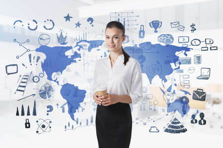 Young businesswoman with coffee standing in blurry office with double exposure of world map and business icons. Toned image.の写真素材