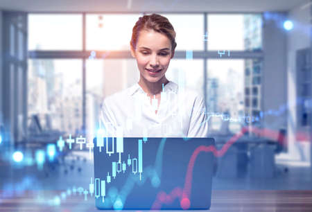 Young businesswoman using laptop in blurry office with double exposure of graphs. Toned image