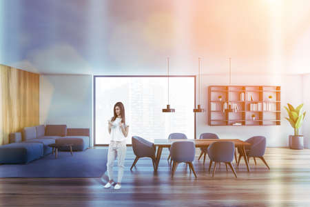 Young woman standing in modern living room with white and wooden walls, wooden floor, sofa and...