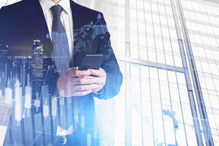 Unrecognizable businessman standing with smarphone in blurry city with double exposure of graphs....