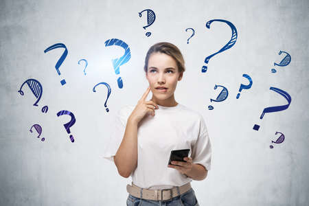 Young woman with smartphone standing near concrete wall with question marks drawn on itの写真素材