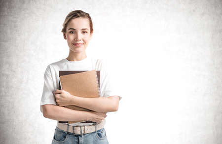 Portrait of smiling young woman holding books near concrete wall. Concept of education. Mock upの写真素材