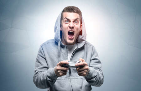 Portrait of young man in hoodie shouting and holding joypad near gray wall. Concept of gaming and...