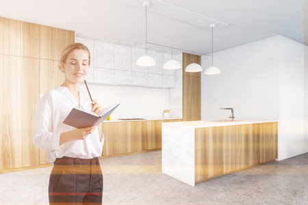 Young businesswoman standing in modern kitchen corner with wooden and white walls, concrete floor and bar. Toned imageの写真素材