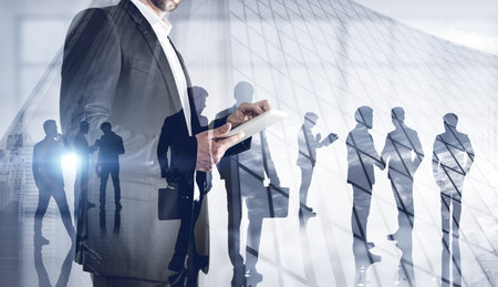 Unrecognizable businessman using tablet in blurry office with double exposure of his business team. Toned imageの写真素材