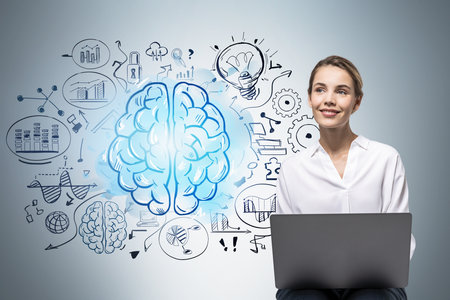 Smiling young woman with laptop sitting near gray wall with brain sketch drawn on it. Concept of...