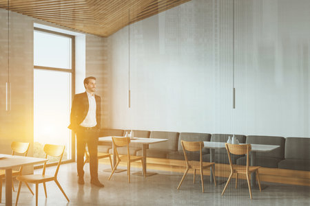 Young European businessman standing in modern restaurant with white walls, concrete floor, gray sofa and square tables. Toned image double exposureの写真素材