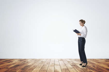 Attractive young woman in formal wear is holding a notebook and pondering how to optimize business process to boost sales of the company. Full length business woman. Wooden parquet floor, concrete wallの写真素材