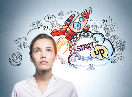 Attractive business woman looking up and pondering about start up and how to succeed on it. Concept of success and new business project launch. Colorful rocket sketch drawn on wallの写真素材