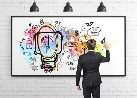 Rear view of a businessman wearing a black suit drawing a large idea project on a white board hanging on a concrete wall in a room. Concept of planning and managementの写真素材