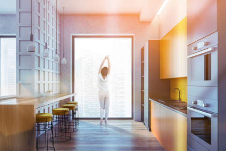 Kitchen set and dining table with bar chairs, parquet floor, woman with city view. Open space dining room with sink and oven, lens flare toned imageの写真素材