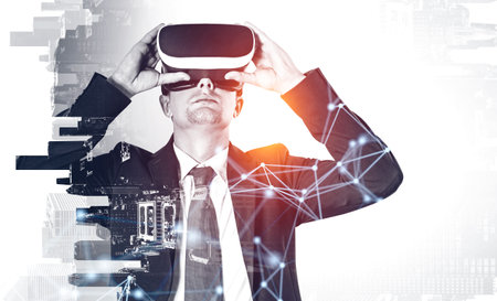 Young handsome businessman in suit holding VR glasses. Double exposure, skyscrapers on background and some digital interconnections and big data. Concept of corporate network and virtual reality.の写真素材