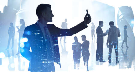Businessman with electronic device, business buildings on background with silhouettes of corporate team. Concept of international communication and partnershipの写真素材