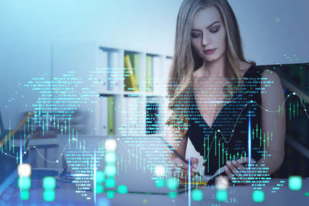 Office woman laptop on table, writing in notebook, working in office interior with double exposure of world map and graph dynamics, candlesticks rising. Concept of stock market and financeの写真素材