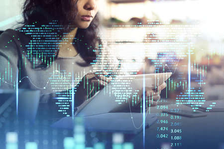 Office woman with tablet in hands, working in blurry office background with double exposure of world map and graph dynamics, candlesticks rising. Concept of stock market and financeの写真素材
