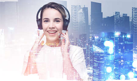 Woman in t-shirt with happy smile wearing headphones, double exposure of office buildings with night lights. Concept of business education and studentの写真素材