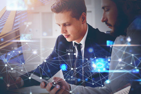 Two businessmen wearing formal suit are working together, holding a smart phone and messaging with their colleagues. Office and skyscraper in the background. Concept of digital technology in businessの写真素材