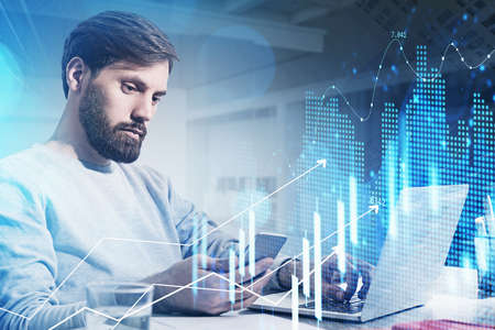 Trader with beard wearing white sweater is sitting at workplace inside the office, holding a smart...