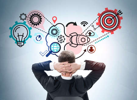 Businessman wearing formal suit is looking at sketch with icons of light bulb, cogwheel, launched rocket, dollar, data cloud, human resources, geolocation. Concept of generation creative business ideaの写真素材