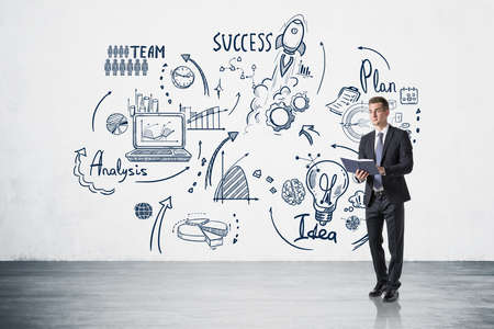 Office man in suit with notebook in hands, standing near business strategy plan on white wall, concrete floor. Doodle drawing with arrows, graphs and network icons. Concept of business ideaの写真素材