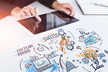 Businesswoman working on touchscreen tablet device, colorful doodle icons and lightbulb. Drawing with arrows, rocket, pie chart and network sketch icons. Concept of fresh idea and business planの写真素材