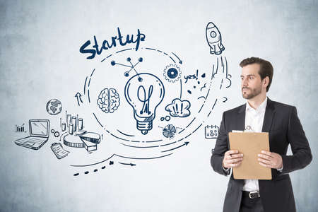 Office man with beard in suit with business files in hands, doodle icons and light bulb on gray concrete wall. Drawing with arrows, pie chart and network icons. Concept of new idea and planの写真素材