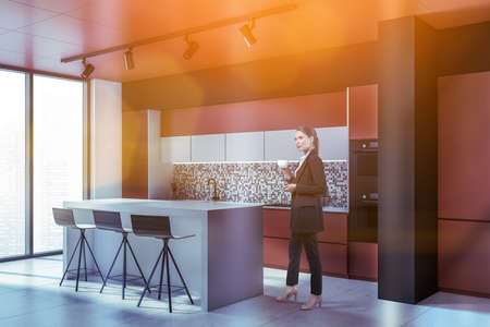 Woman at kitchen room with furniture and panoramic window, lens flare. Businesswoman drinking coffee...