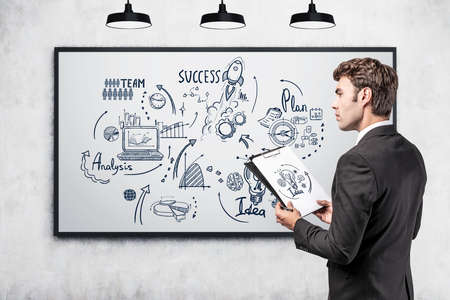 Office man in suit with business files in hands, business strategy plan in a frame on gray wall. Doodle drawing with arrows, graphs and network icons. Concept of analysisの写真素材
