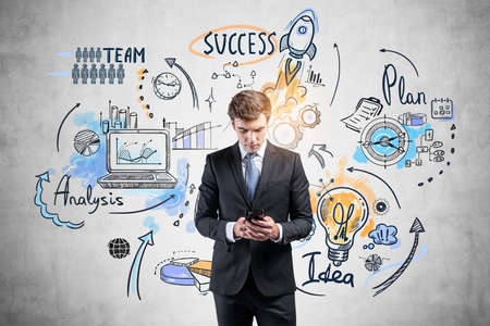 Office man in suit with smartphone in hands, colorful business strategy plan on gray wall, drawing with arrows, graphs and network icons. Concept of business idea and technologyの写真素材