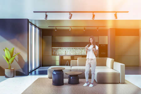 Attractive young woman with phone in modern living room with kitchen and dining area, lens flare. Sink, cupboard with kitchenware and panoramic windowsの写真素材