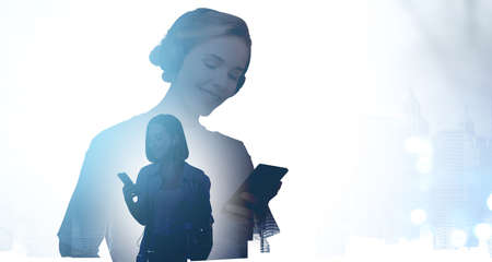Two young women students in casual clothes, smiling typing on the smartphones, wearing headphones, double exposure. Concept of communication and technologyの写真素材