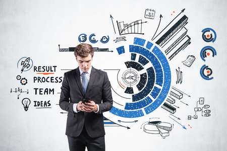 Office man in suit typing in smartphone. Background of colorful doodle pie chart and bars, business plan on concrete wall. Concept of technology and business processの写真素材